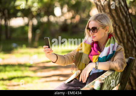 Blonde Frau ein Selbstporträt mit Smartphone auf Bank im Park sitzen zu tun. Stockfoto