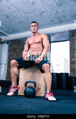 Muskulöser Mann auf Fit Box sitzen und lesen für Training mit Wasserkocher Ball im Fitness-Studio. Blick in die Kamera Stockfoto