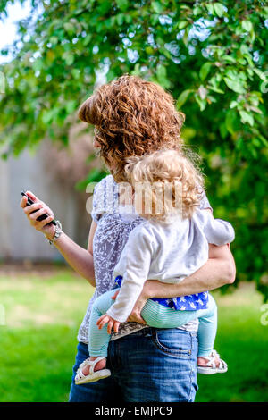 Eine 50 Jahre alte kaukasische Großmutter hält 14 Monate alte Enkelin beim Telefonieren Handy im Freien. USA Stockfoto