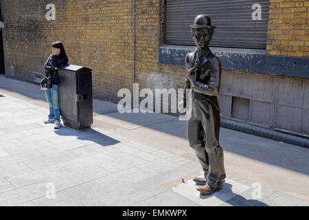 Eine Statue von Charlie Chaplin in der Nähe von Leicester Square, London, England, Vereinigtes Königreich mit Frau in der Nähe am Telefon zu sprechen. Stockfoto