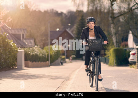 Junge Geschäftsfrau, die Fahrt zur Arbeit mit dem Fahrrad entlang einer Wohnstraße in ihrem stilvollen schlaff Anzug und Sicherheit Helm in einer eco Stockfoto