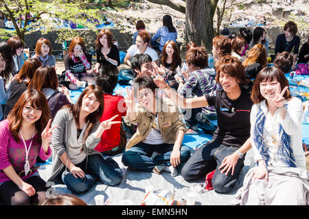 Kirschblütenparty, Japan. Große Gruppe von Teenagern, die auf blauem Plastikblech mit Essen und Getränken sitzen und sich des Betrachters bewusst sind, so lächelnd und gestisch. Stockfoto