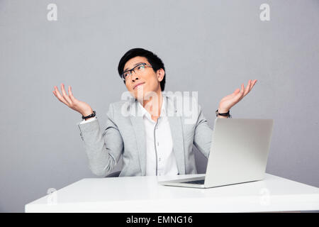Junger Geschäftsmann mit Laptop am Tisch sitzen und Achselzucken auf grauem Hintergrund. Ich weiß es nicht. Stockfoto