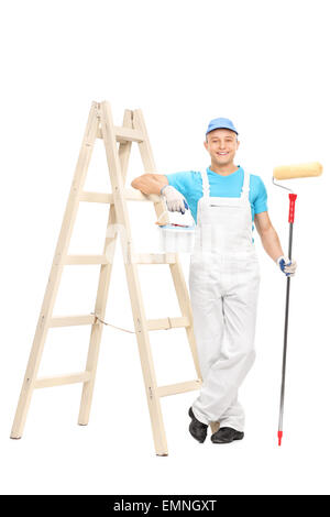 Junge männliche Anstreicher in einen weißen sauberen Overall hält einen Farbroller und stützte sich auf eine Holzleiter Stockfoto
