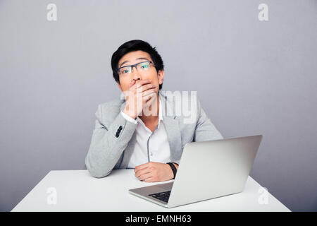 Erstaunt, Geschäftsmann, sitzen am Tisch mit Laptop über grauen Hintergrund und Blick in die Kamera. Sein Mund Stockfoto