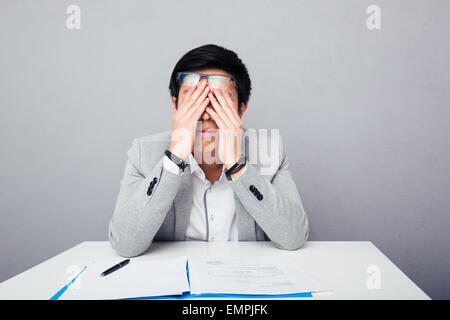 Junge asiatische Geschäftsmann am Tisch sitzen und rieb seine Augen über grauen Hintergrund Stockfoto