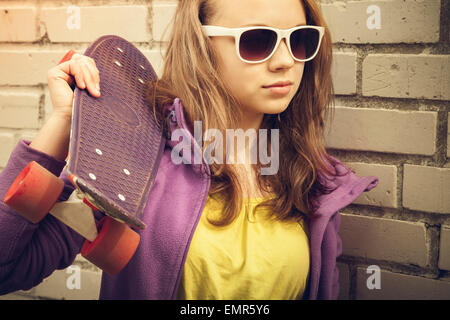 Blonde Teenager-Mädchen in Sonnenbrille hält Skateboard in der Nähe von grau urban Mauer, Vintage warmer Tonwertkorrektur, alten Stil Foto Stockfoto