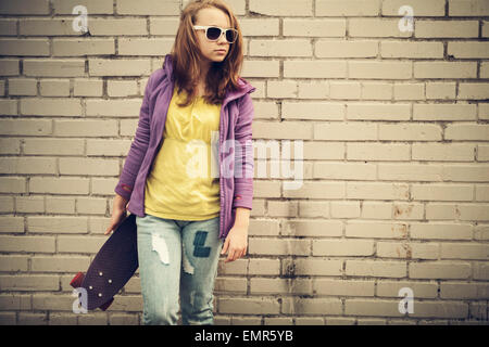 Blonde Teenager-Mädchen in Jeans und eine Sonnenbrille hält Skateboard in der Nähe von grau urban Mauer, Vintage Tonwertkorrektur, alten Stil Stockfoto