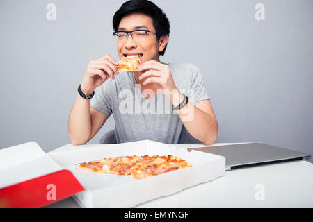 Junge asiatische Mann isst Pizza im Büro über grauen Hintergrund Stockfoto
