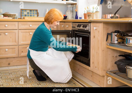 Blondes Mittelalter Frau mit Schürze, einen Kuchen für Snacks in einem Ofen in der Küche zu Hause. Stockfoto