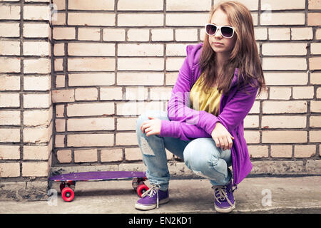 Blonde Teenager-Mädchen in eine Sonnenbrille mit Skateboard sitzt in der Nähe von urban Mauer, Foto mit Retro-Tonwertkorrektur-Effekt, instagr Stockfoto