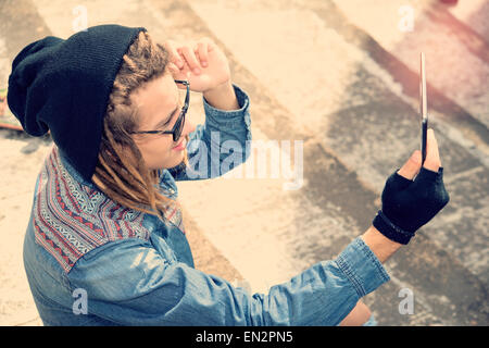 hübscher Kerl mit Dreadlocks sitzt auf der Treppe nehmen Selfie mit Tablet warm Filter angewendet Stockfoto