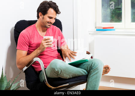 Junger Mann in Freizeitkleidung auf schwarzen Stuhl sitzend und ein Buch lesen und hält ein Glas des Getränks. Stockfoto