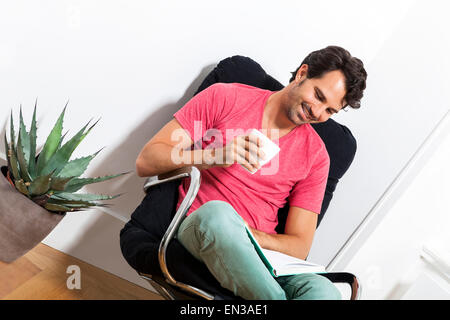 Junger Mann in Freizeitkleidung auf schwarzen Stuhl sitzend und ein Buch lesen und hält ein Glas des Getränks. Stockfoto