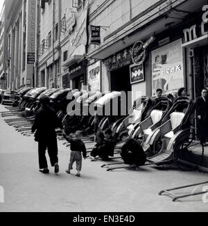 Historisches Bild der 1950er Jahre, Hongkong, in einer schrägen Seitenstraße, eine Reihe gezogener Rikschas oder Passagierwagen. Die meisten Rikschas haben ihre Hauben nach oben, aber drei haben ihre Hauben nach unten, um zu zeigen, dass sie gemietet werden können. Ihre Betreiber spielen ein Spiel auf der Straße. Rikschas sind seit Jahrhunderten eine beliebte Form des Taxiverkehrs, ihr Name stammt aus den japanischen für den menschlichen Transport. Büros der South China Morning Post Zeitung sind zu sehen, und die Rikschas sind vor dem Blue Theatre und dem Nachtclub geparkt. Stockfoto