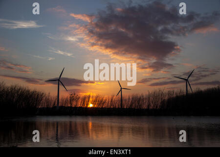 Windenergieanlagen bei Sonnenuntergang mit Reflexionen in einem See in den Vordergrund und bunten Wolken hinter. Stockfoto