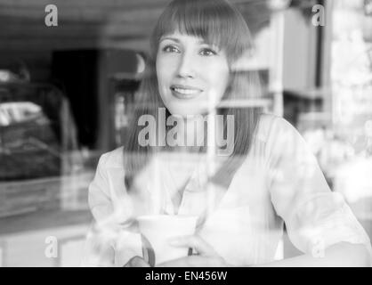 Kaukasische Mädchen im Kaffeehaus am Sommermorgen. Hübsches Mädchen mit Tasse Kaffee hinter Glas. Schwarz / weiß Bild Stockfoto