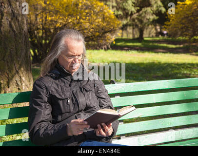 Behaartes älterer Mann in einer schwarzen Jacke und Brille mit einem Buch sitzen auf einer Bank im Stadtpark, An sonnigen Frühlingstag Stockfoto