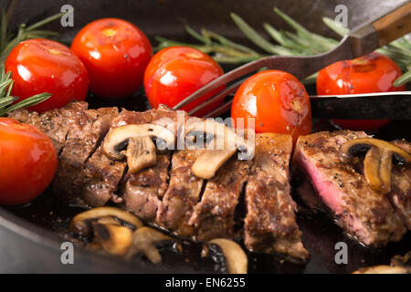 Serie: Strip Loin Steak in Gusseisen Pfanne Grillen: Steak gekocht und in Scheiben geschnitten - mit gegrillten Tomaten und Mushr Stockfoto