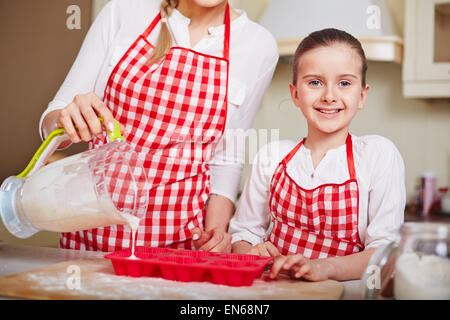 Entzückende Mädchen Blick in die Kamera, während ihre Mutter in Muffin-Formen mit flüssigen Teig füllen Stockfoto