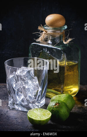 Glas mit Eiswürfeln und Flasche Tequila, serviert mit in Scheiben geschnittenen Limetten auf schwarzem Hintergrund Stockfoto