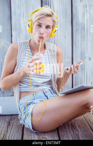 Hübsche blonde Frau mit ihrem Handy Musik hören und trinken Orangensaft Stockfoto