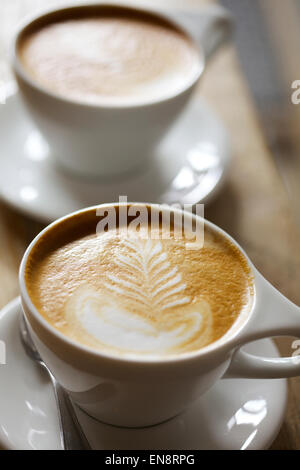 Zwei Cappuccino mit Blattmuster in den Schaum in weißen Tassen mit Untertassen auf einem Holztisch sitzend, hat Hintergrund weichzeichnen. Stockfoto