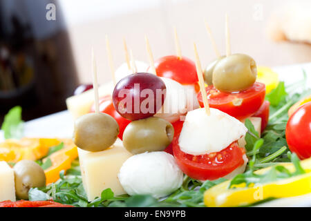 Fingerfood: Spieße mit Käse, Oliven, Trauben, Tomaten auf einem Teller mit frischem Rucola und Paprika, Stockfoto