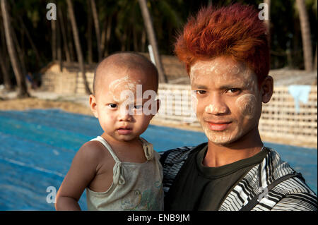 Junge burmesische Fischer mit funky gefärbtes Haar mit seinem Baby am Ngapali Strand mit blauen Fischernetze hinter ihm Stockfoto