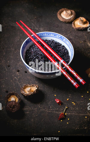 Porzellanschüssel mit ungekochtem Reis schwarz, serviert mit trockenen Shiitake-Pilzen und roten Stäbchen über dunklen Tisch. Stockfoto