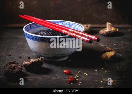 Porzellanschüssel mit ungekochtem Reis schwarz, serviert mit trockenen Shiitake-Pilzen und roten Stäbchen über dunklen Tisch. Stockfoto