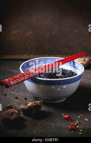 Porzellanschüssel mit ungekochtem Reis schwarz, serviert mit trockenen Shiitake-Pilzen und roten Stäbchen über dunklen Tisch. Stockfoto