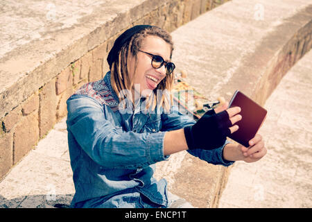 hübscher Kerl mit Dreadlocks sitzt auf der Treppe nehmen Selfie mit Tablet warm Filter angewendet Stockfoto