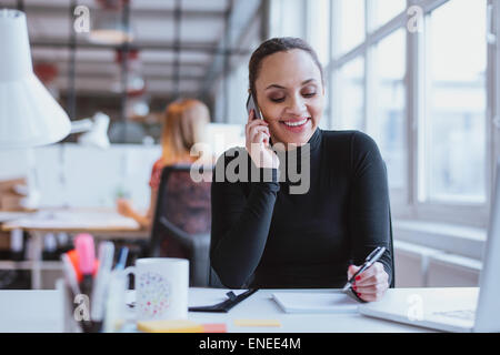 Glückliche junge Frau sitzt an ihrem Schreibtisch arbeiten und einen Anruf zu beantworten. Stockfoto