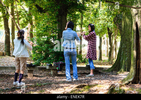 Drei japanische Teenager-Frauen, die in einem Halbkreis an einer Bank in einer Lichtung in einigen sonnenbeschienenen Bäumen stehen und Flöten spielen. Frauen sind lässig gekleidet. Stockfoto