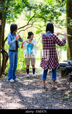 Drei japanische Teenager-Frauen, die in einem Halbkreis an einer Bank in einer Lichtung in einigen sonnenbeschienenen Bäumen stehen und Flöten spielen. Frauen sind lässig gekleidet. Stockfoto