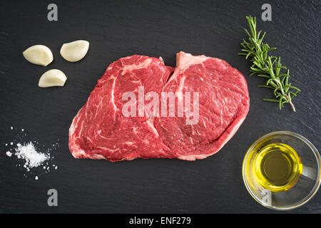 Frische ungekochte Rib-Eye-Steak mit Knoblauch, Salz, Olivenöl und Rosmarin auf schwarzem Hintergrund Stockfoto