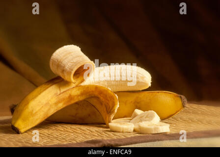 2 reife Bananen, Bambus Tischläufer auferlegt. eine Banane ist teilweise geschält, mit einigen Scheiben vorne gedreht im Studio auf eine gree Stockfoto
