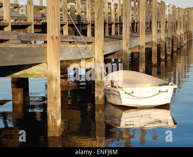 Eine kleine, weiße Ruderboot ist neben einer langen hölzernen Pier in der späten Nachmittagssonne, wirft seine Reflexion auf die Wate gefesselt Stockfoto