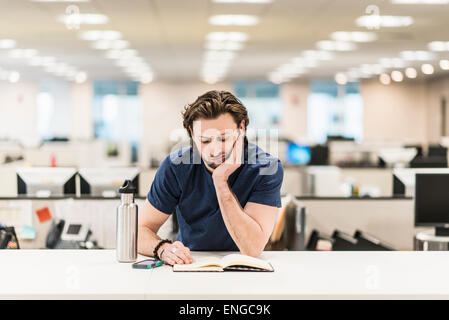 Ein Mann stützte sich auf seine Ellbogen und mit Blick auf ein offenes Buch auf einem Schreibtisch. Stockfoto