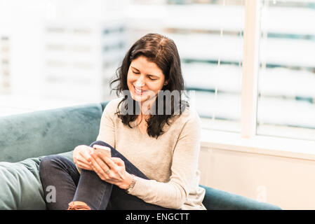 Eine Frau sitzend mit den Füßen oben auf einem Sofa, Blick auf ihr Smartphone. Stockfoto