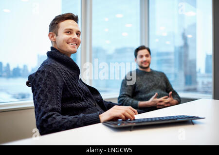 Zwei Männer in einem Büro sitzen an einem Schreibtisch im Chat und lächelnd. Stockfoto