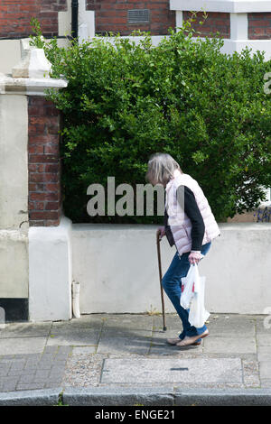 ältere Dame mit gebeugtem Rücken möglicherweise von Osteoporose zu Fuß entlang der Fahrbahn mit ihr Einkaufen Stockfoto