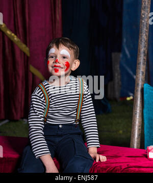 Niedlicher fröhlicher Junge mit Clown Make-up sitzt auf einer Bühne vor einer Vertretung während des Tragens gestreiftes Langarm-Hemd und Hosenträger Stockfoto