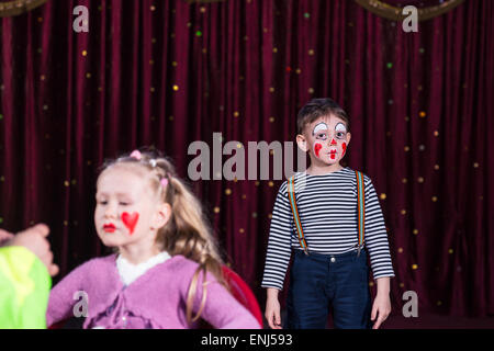 Junge verkleidet als Clown tragen Make-up und gestreiftes Hemd Mädchen machen bis auf Gesicht, das ich mit roten Vorhang auf der Bühne stehen Stockfoto