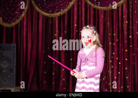 Lächelnden blonden Mädchen tragen Clown Make Up und gestreiften Kleid Standing on Stage Holding über große rosa Kamm vor Rot Curtai Stockfoto