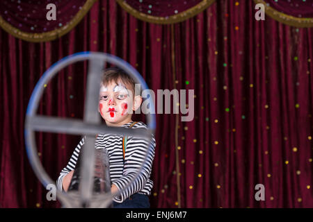 Kleiner Junge, als tragen Make Up Clown verkleidet und gestreiftes Hemd suchen ernsthafte und eingerahmt in Eisen-Visier und auf der Bühne stehen Stockfoto