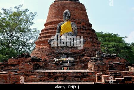 Ayutthaya, Thailand: Einen riesigen Stein sitzende Buddha mit einer orangefarbenen Schärpe vor einem Ziegelstein glockenförmigen Chedi Stockfoto