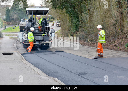 Männer, die bei Straßenarbeiten arbeiten Resurfacing & Reparaturen mit mechanischen Asphalt Spreitmaschine tragen hohe Sichtbarkeit Jacken & Schutzhelme Essex England UK Stockfoto