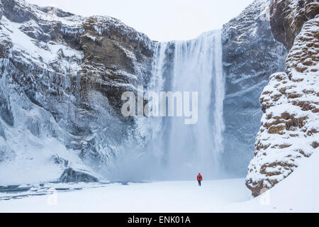 Eine Person im roten Jacke Wandern im Schnee in Richtung Skogafoss Wasserfall im Winter, Skogar, South Island, Island Stockfoto